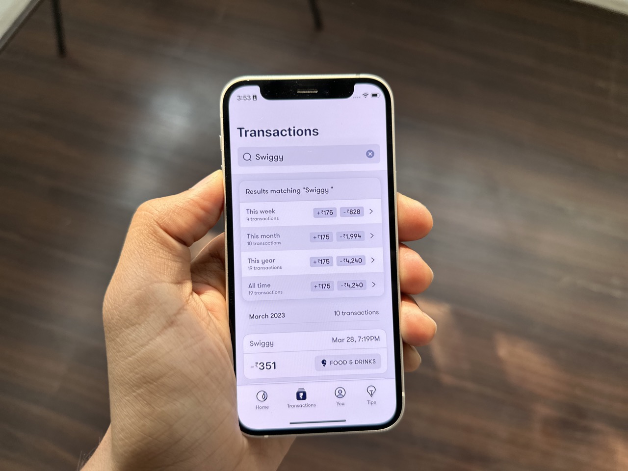 A person holding the iphone with Fold app open. In the app - "swiggy" is in transaction search field and in results there is a summary which shows transactions in  this week, this month, this year, and all time with amounts. 